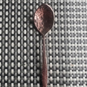 May include: A copper-colored spoon with a hammered texture on the bowl and handle. The spoon is centered against a black and gray woven background, creating a contrast in textures and colors. The spoon's design suggests a rustic or handcrafted aesthetic.