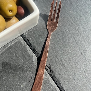 May include: A copper-colored olive fork with three tines, set against a dark gray slate surface. The fork has a hammered texture and a long, rectangular handle. A white rectangular dish filled with green and red olives is in the background.