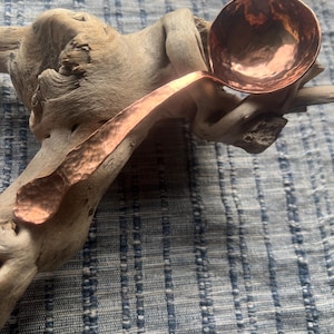 May include: A handcrafted copper ladle with a hammered texture, resting on a piece of driftwood. The ladle features a rounded bowl and a long, curved handle. The copper has a warm, reddish-brown hue, contrasting with the neutral tones of the wood.