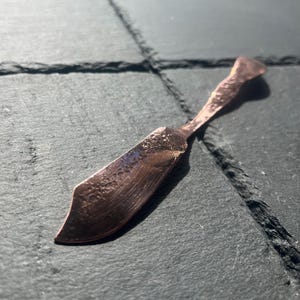 May include: A handcrafted copper butter knife with a textured surface. The knife has a curved blade and a handle with a hammered appearance. The copper color gives it a rustic and elegant look.