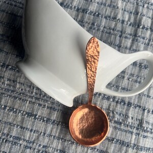 May include: A copper-colored measuring spoon with a hammered texture rests near a white ceramic pitcher. The spoon has a round bowl and a long handle. The background is a blue and white striped fabric.