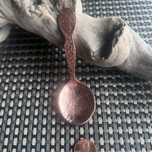 May include: A copper-colored spoon with a textured surface. The spoon is placed on a patterned surface next to a penny. The spoon's handle has a hammered design. The spoon is approximately 6 inches long.