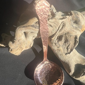 May include: A copper spoon with a hammered texture. The spoon has a long handle and a rounded bowl. The copper has a reddish-brown hue and reflects light. The spoon is displayed against a backdrop of driftwood.