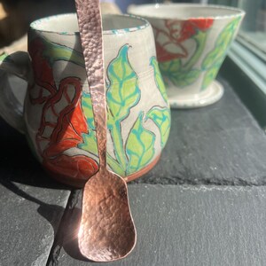 May include: A copper spoon with a hammered texture rests in front of a ceramic mug and bowl. The mug and bowl are decorated with red and green floral designs on a white background. The spoon is approximately 7 inches long.