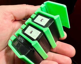 Peut inclure: Un porte-cartouches d'encre d'imprimante en plastique vert. Le porte-cartouches a des emplacements pour trois cartouches et un couvercle à charnière. Les cartouches sont étiquetées avec les numéros "3" et "5".