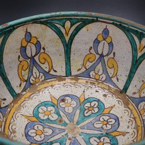 May include: A ceramic bowl with a white background and intricate blue, green, and yellow floral designs. The bowl has a worn, antique look.