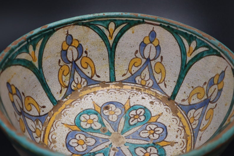 May include: A ceramic bowl with a white background and intricate blue, green, and yellow floral designs. The bowl has a worn, antique look.