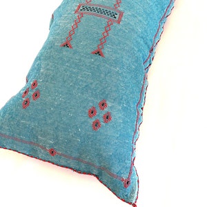 Pu&ograve; includere: Un cuscino blu e rosso ricamato con un motivo geometrico. Il cuscino &egrave; realizzato in un tessuto morbido e testurizzato e ha un fiocco su un angolo.