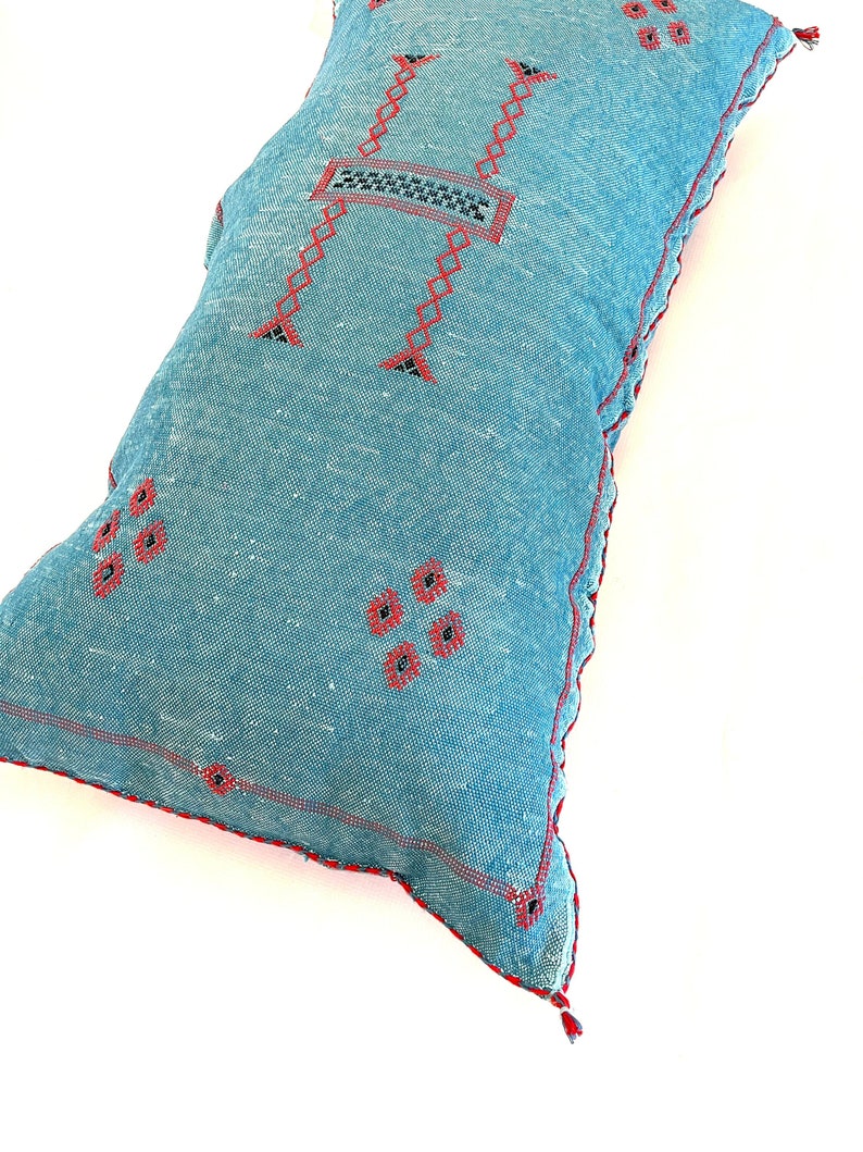 Pu&ograve; includere: Un cuscino blu e rosso ricamato con un motivo geometrico. Il cuscino &egrave; realizzato in un tessuto morbido e testurizzato e ha un fiocco su un angolo.