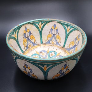 May include: A white ceramic bowl with a green, blue, and yellow floral design. The bowl is decorated with intricate patterns and has a slightly rounded shape.