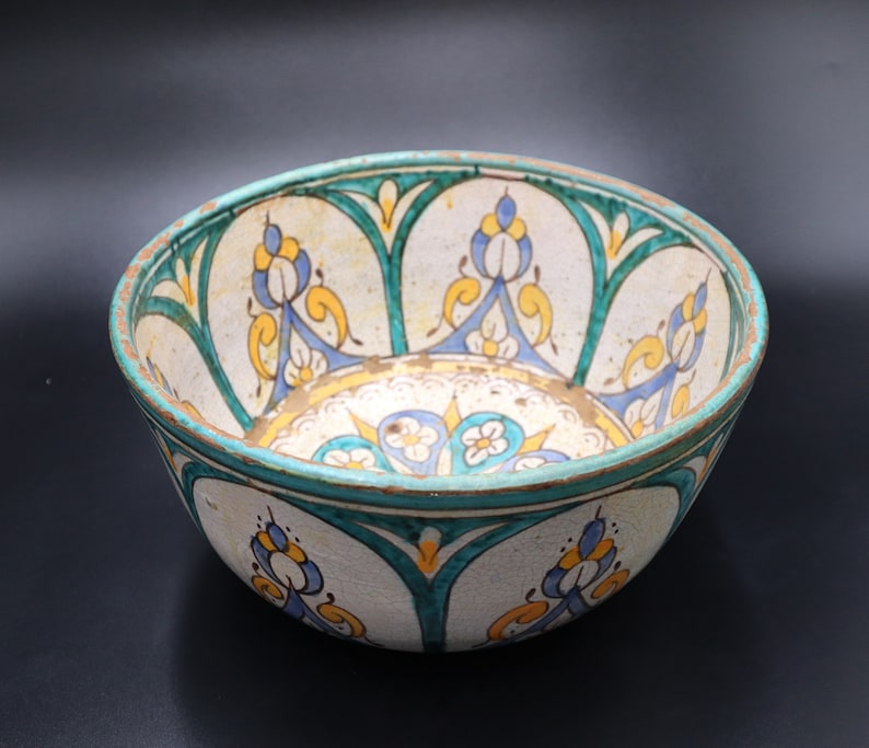 May include: A white ceramic bowl with a green, blue, and yellow floral design. The bowl is decorated with intricate patterns and has a slightly rounded shape.