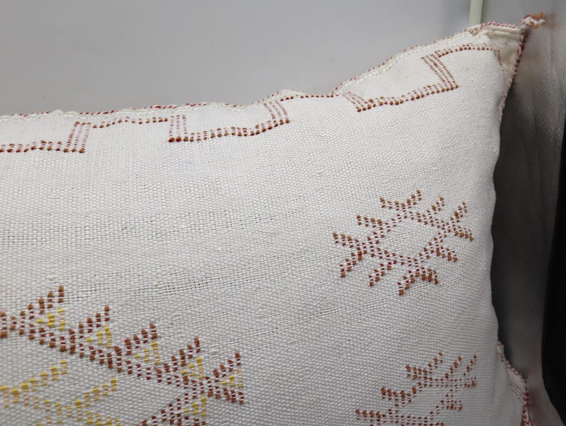May include: White throw pillow with a geometric pattern in brown and yellow thread. The pattern is a repeating design of stylized stars.