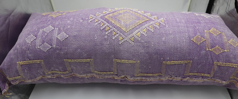 Puede incluir: Un coj&iacute;n de tiro de color morado y dorado con un dise&ntilde;o geom&eacute;trico. El coj&iacute;n est&aacute; hecho de una tela suave y tejida.