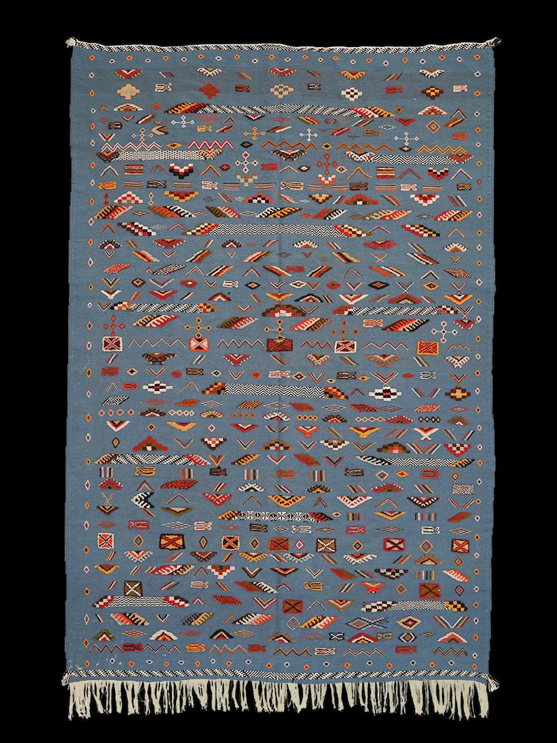 May include: A rectangular, blue rug with a complex geometric pattern. The design incorporates various shapes and colors, including red, orange, yellow, and white, creating a visually rich textile. The rug has a fringe along the bottom edge.