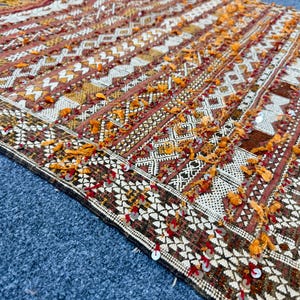 K&ouml;nnte beinhalten: Nahaufnahme eines dekorativen Textils mit geometrischem Muster. Der Teppich weist eine Mischung aus Braun-, Wei&szlig;- und Oranget&ouml;nen mit aufwendigen Designs und Fransen auf. Das Textil wird wahrscheinlich als Teppich oder Wandbehang verwendet.