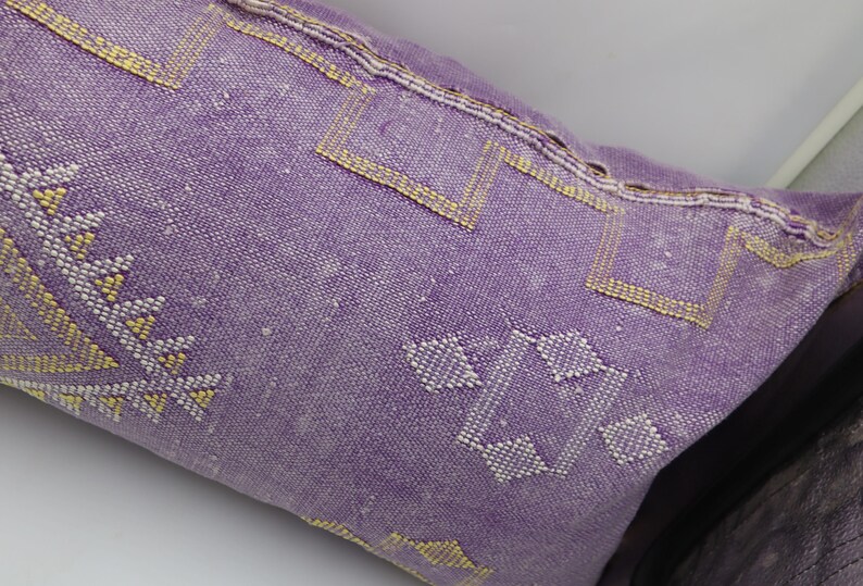 Puede incluir: Una almohada morada con un patr&oacute;n geom&eacute;trico en hilo blanco y amarillo. La almohada est&aacute; hecha de una tela suave y tejida.