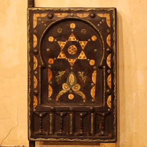 以下が含まれることがあります： 複雑な象眼細工のデザインが施された濃い茶色の木製パネル。パネルの中央にはダビデの星のデザインがあり、クリーム色、金色、ティール色の花柄と幾何学模様で囲まれています。パネルは長方形で、上部が湾曲しており、装飾的なトリムが付いています。