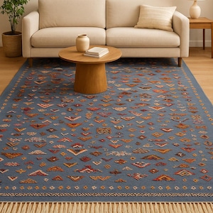 May include: A blue area rug with a geometric pattern in shades of orange, red, and cream. The rug has a fringe border and is placed in a living room setting with a beige sofa and wooden furniture.