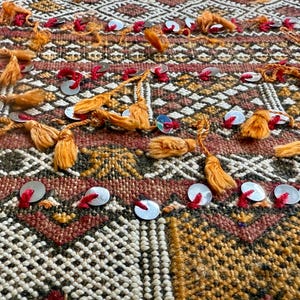 K&ouml;nnte beinhalten: Nahaufnahme eines strukturierten Textils mit einem geometrischen Muster in Braun-, Orange- und Cremet&ouml;nen. Der Stoff ist mit orangefarbenen Quasten, roten N&auml;hten und kleinen silbernen Pailletten verziert, was ein dekoratives Design ergibt.