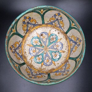 May include: A white ceramic bowl with a blue, green, and yellow floral design. The bowl has a scalloped edge and a raised center.