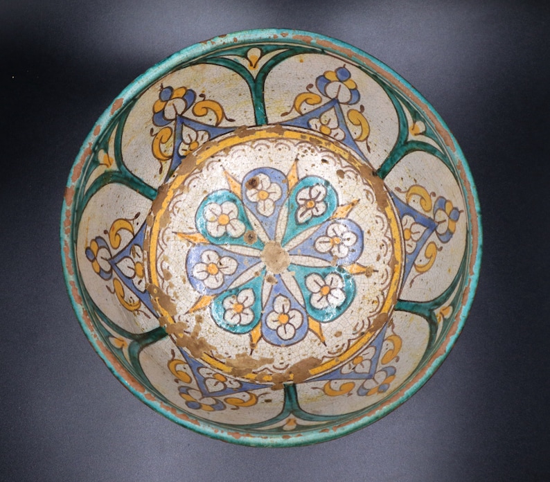 May include: A white ceramic bowl with a blue, green, and yellow floral design. The bowl has a scalloped edge and a raised center.