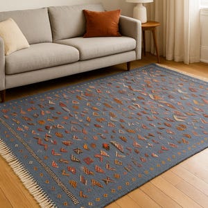 May include: A blue area rug with a geometric pattern in shades of orange, red, and cream. The rug has a fringe on one end and is placed on a hardwood floor. A gray sofa and pillows are in the background.