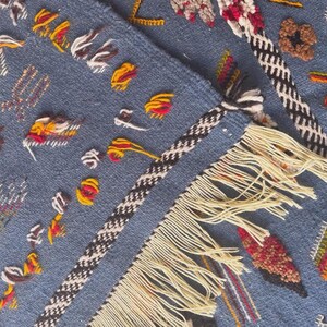 May include: Close-up of a blue textile with intricate embroidery. The design features colorful stitched details in red, yellow, and white, along with a black and white braided border and cream-colored fringe. The textile has a textured appearance.