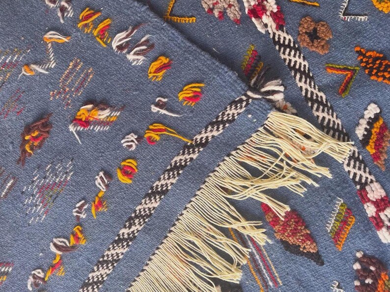 May include: Close-up of a blue textile with intricate embroidery. The design features colorful stitched details in red, yellow, and white, along with a black and white braided border and cream-colored fringe. The textile has a textured appearance.