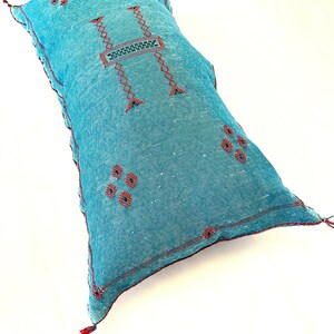 Pu&ograve; includere: Un cuscino blu turchese con cuciture rosse e dettagli ricamati. Il cuscino ha un fiocco su un angolo.
