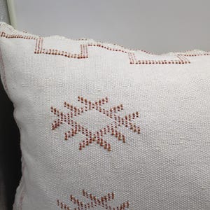 May include: White throw pillow with a geometric pattern in shades of brown. The pillow has a woven texture and a fringed edge.