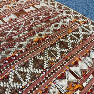 K&ouml;nnte beinhalten: Nahaufnahme eines gewebten Textils mit geometrischem Muster. Das Design zeigt Rauten- und Dreiecksformen in Braun-, Rot- und Cremet&ouml;nen, akzentuiert mit orangefarbenen und silbernen Pailletten. Das Textil hat ein strukturiertes Aussehen.