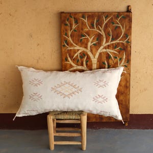 May include: A white throw pillow with a geometric pattern in shades of pink and orange. The pillow is sitting on a wooden stool in front of a wooden wall with a carved tree design.