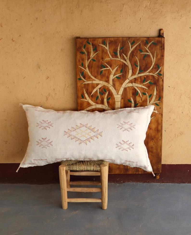 May include: A white throw pillow with a geometric pattern in shades of pink and orange. The pillow is sitting on a wooden stool in front of a wooden wall with a carved tree design.