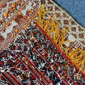 K&ouml;nnte beinhalten: Nahaufnahme eines Teils eines gewebten Teppichs mit komplizierten Mustern. Der Teppich weist eine Mischung aus Farben auf, darunter Rot, Braun und Creme, mit Akzenten aus gelben Fransen. Das Design umfasst geometrische Formen und detaillierte Bord&uuml;ren.
