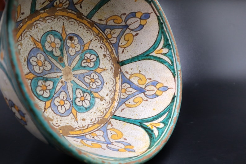 May include: A ceramic bowl with a white background and a colorful floral design. The bowl has a green rim and a blue and yellow floral pattern.