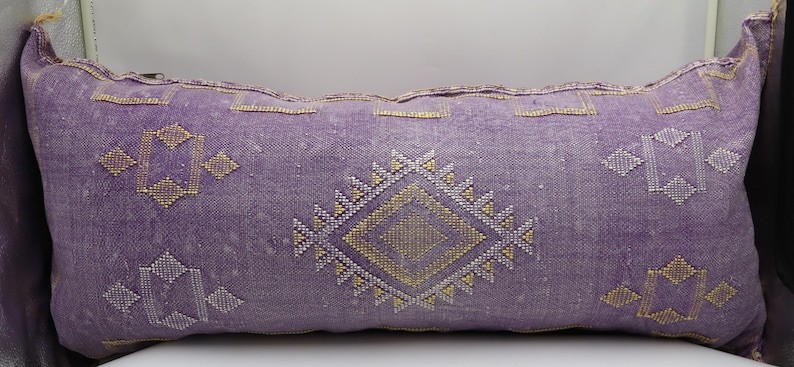 Puede incluir: Una almohada de tiro de color morado y dorado con un dise&ntilde;o geom&eacute;trico. La almohada est&aacute; hecha de una tela suave y tejida y tiene un aspecto desgastado sutil.