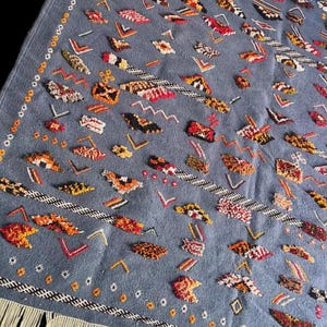May include: A blue rug with a detailed pattern of various shapes in red, orange, yellow, and white. The rug has a fringe along one edge and a decorative border.