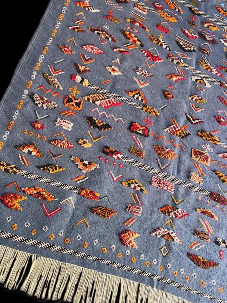 May include: A blue rug with a detailed pattern of various shapes in red, orange, yellow, and white. The rug has a fringe along one edge and a decorative border.
