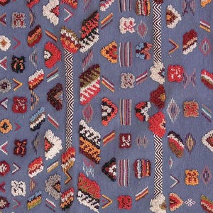 May include: Close-up of a blue textile with a repeating pattern of colorful, textured shapes. The shapes include triangles, rectangles, and other geometric forms in shades of red, orange, white, and brown. Vertical stripes with a zigzag pattern are also present.
