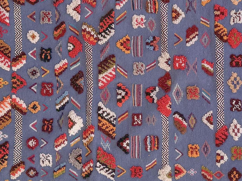May include: Close-up of a blue textile with a repeating pattern of colorful, textured shapes. The shapes include triangles, rectangles, and other geometric forms in shades of red, orange, white, and brown. Vertical stripes with a zigzag pattern are also present.