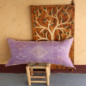 Puede incluir: Una almohada larga y rectangular de color morado con un patr&oacute;n geom&eacute;trico blanco y dorado. La almohada est&aacute; sentada en un taburete de madera frente a una pared de madera con un dise&ntilde;o de &aacute;rbol tallado.