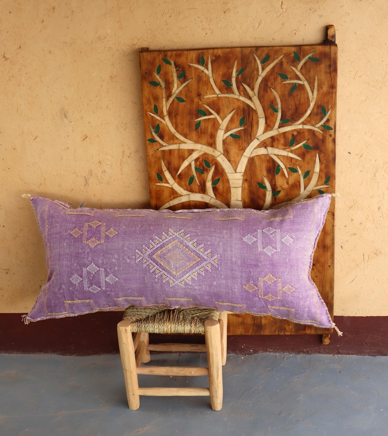 Puede incluir: Una almohada larga y rectangular de color morado con un patr&oacute;n geom&eacute;trico blanco y dorado. La almohada est&aacute; sentada en un taburete de madera frente a una pared de madera con un dise&ntilde;o de &aacute;rbol tallado.