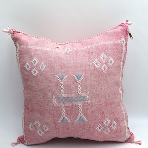 May include: A pink cushion with a white and blue geometric pattern. The cushion has a zipper closure and tassels on the corners.