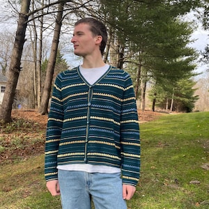 Op de afbeelding: Een groene cardigan met gele en blauwe horizontale strepen. De cardigan heeft een V-hals en is dichtgeknoopt. De persoon draagt een wit t-shirt en een blauwe jeans.