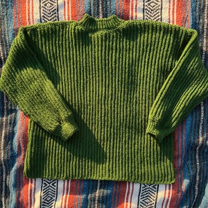 K&ouml;nnte beinhalten: Ein gr&uuml;ner Strickpullover mit Stehkragen und langen &Auml;rmeln. Der Pullover ist aus einem dicken Garn gefertigt und hat eine gerippte Struktur.