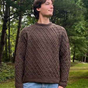 Puede incluir: Un suéter de manga larga de color marrón oscuro con un patrón de diamantes texturizado en la parte delantera y las mangas, cuello redondo y puños y dobladillo acanalados. La prenda se combina con pantalones vaqueros azul claro.