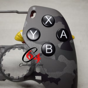 Puede incluir: Primer plano de una placa frontal de mando de videojuego de color gris camuflaje con botones negros etiquetados con las letras X, Y, A y B. La placa frontal está parcialmente unida a una carcasa de mando gris.
