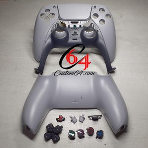 Op de afbeelding: Een gedemonteerde witte PlayStation 5 DualSense-controller met alle knoppen en onderdelen die op een grijs oppervlak zijn gelegd. De controller bestaat uit twee delen, de bovenste helft en de onderste helft. De knoppen zijn allemaal zwart en wit.