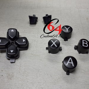 Könnte beinhalten: Ein Satz schwarzer Ersatztasten für einen Videospielcontroller. Die Tasten haben weiße Beschriftungen mit den Buchstaben A, B, X, Y und Richtungspfeilen.