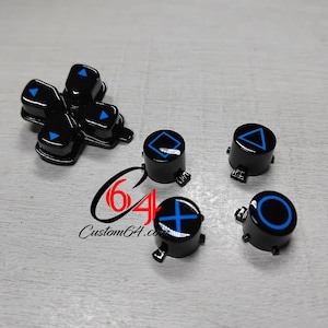 May include: Black replacement buttons for a game controller, including a directional pad and four circular buttons. Each button features a blue symbol. The text "Custom64.com" is also visible.